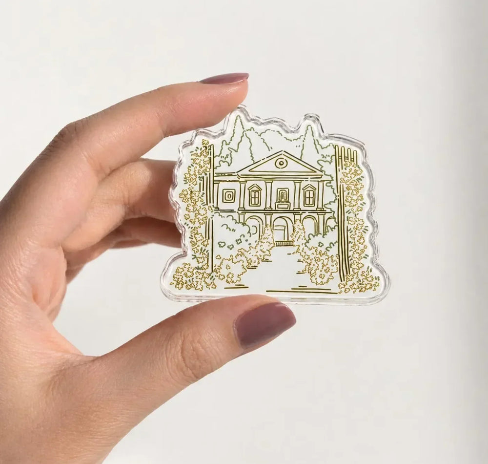 Hand holding a shaped acrylic magnet with a fine-line building illustration and green vines.