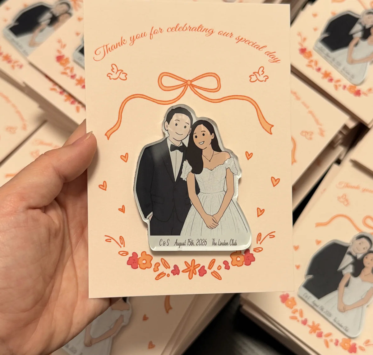 Hand holding a couple illustration acrylic magnet on a blush backing card decorated with a bow and small hearts.