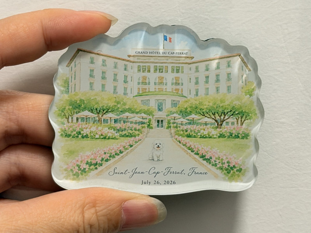 Hand holding a custom acrylic fridge magnet with a pale venue building, green gardens, and a central path.
