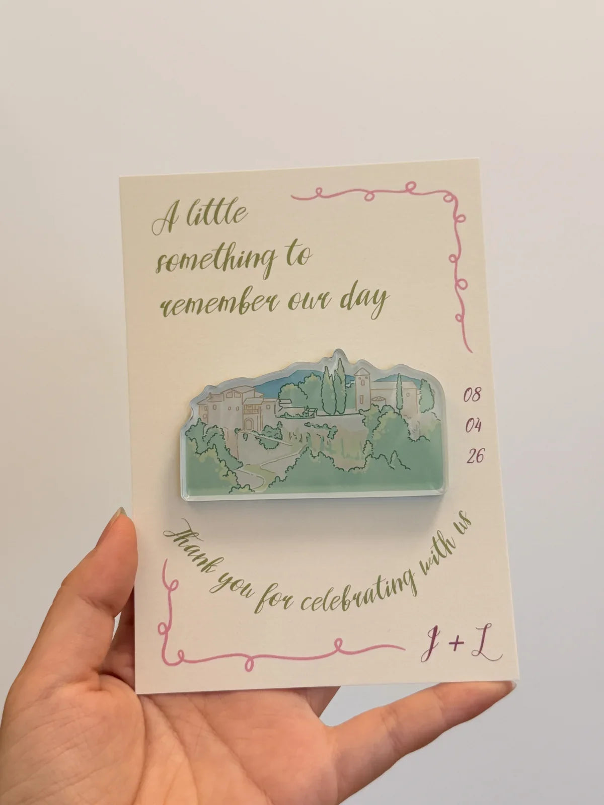 Green illustrated venue magnet on a cream wedding favor card with olive lettering and pink border details