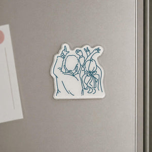 May include: A clear, rectangular magnet with a blue outline illustration of two people with their arms raised. One figure has long hair tied with a bow. The magnet is adhered to a silver refrigerator.
