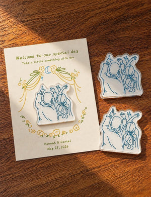 May include: A wedding invitation card with the text "Welcome to our special day" and "Take a little something with you". The card is decorated with floral and moon illustrations. Three clear acrylic wedding favors with a blue line drawing of a couple are also present.